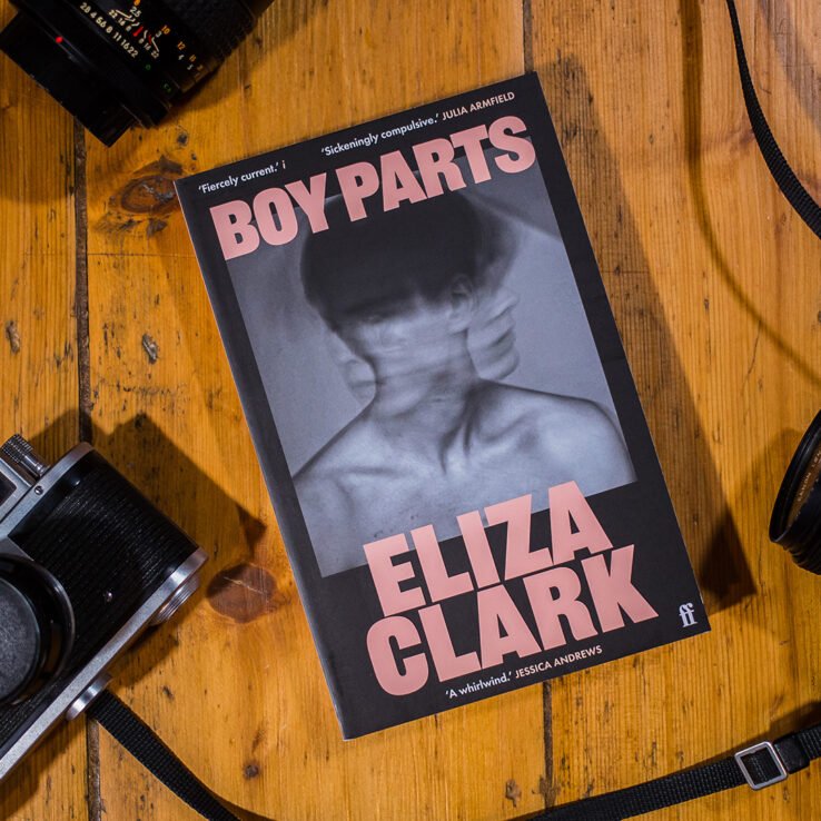 The paperback of Boy Parts by Eliza Clark on a wooden table next to vintage camera